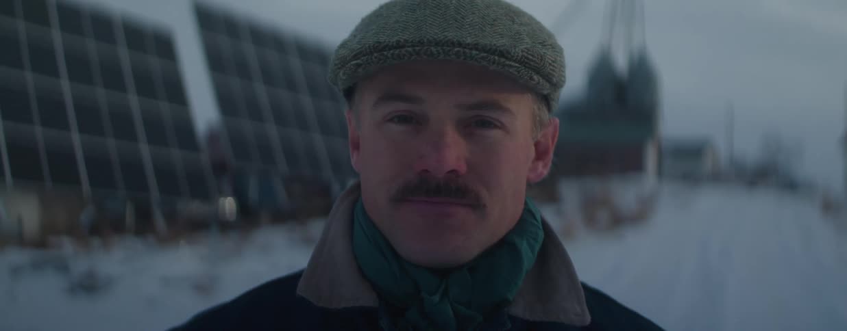 A close-up portrait of a confident man with a mustache and a flat cap looking at the camera in a snowy, rural setting with solar panels in the background