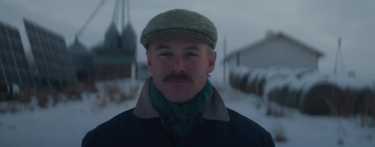 A portrait of a confident farmer with a mustache and cap looking at the camera on a snowy, cold winter day on his farm