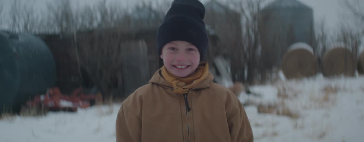 A young boy in a beanie and warm jacket smiles happily while standing outside in the snow on a rural farm