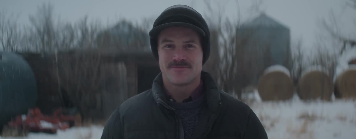 A young farmer with a mustache gives a warm smile on a cold winter day, with his snowy farm and silos in the background