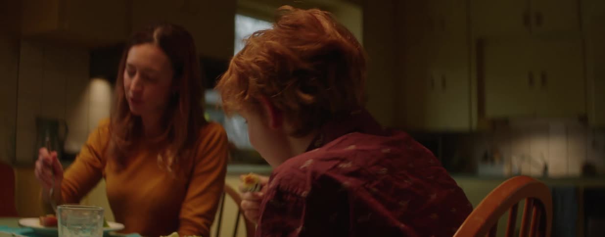 A smiling young boy with red hair enjoys dinner and conversation with his mother at the dining table in their home