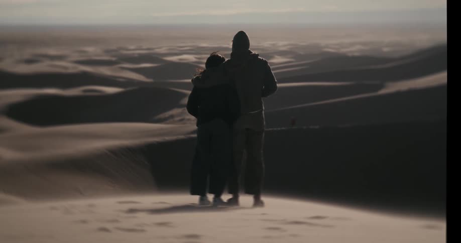A cinematic slow-motion shot shows a couple standing together and embracing while looking out over a vast and windy desert of sand dunes