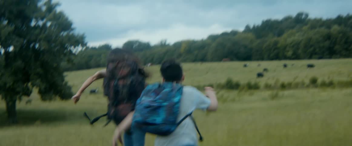 Two survivors with backpacks frantically run across an open field in a suspenseful escape