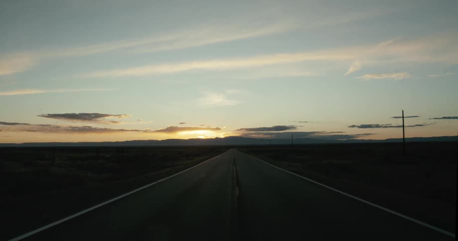 A serene forward-moving shot down the center of a long, empty desert highway toward the setting sun on the horizon