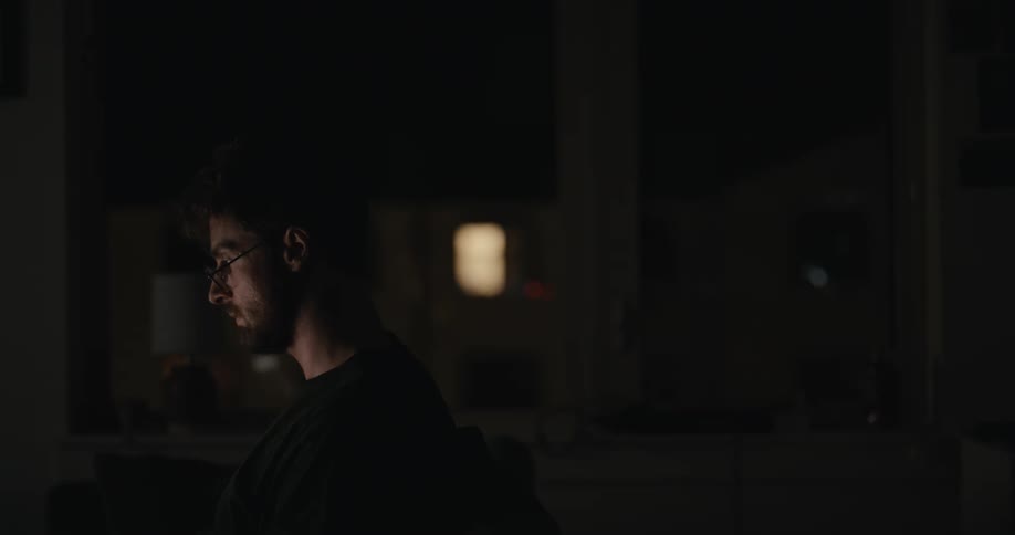Side profile of a young man working late at night in a dark room, his face illuminated by the bright computer screen