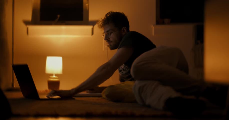 A focused student lying on the floor in a dimly lit room takes notes and works on his laptop late into the night