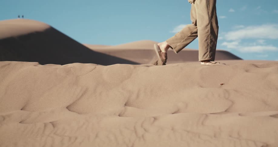 Slow motion close-up of a person's feet in sandals walking across a vast desert sand dune