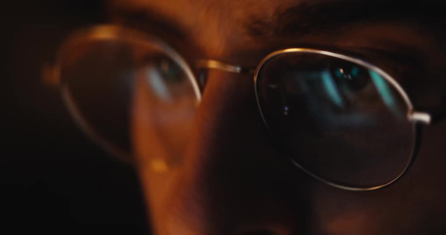 An extreme close-up shot shows a computer screen with scrolling text reflected in a person's glasses in a dark room