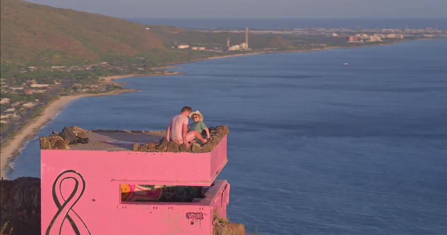 An aerial view reveals a couple sitting atop a pink bunker on a cliff, then pulls back to show the man reading alone against the vast ocean backdrop
