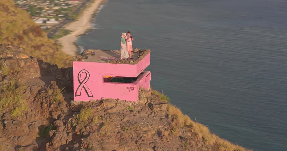 An aerial shot circles around three friends taking a selfie on top of a bright pink bunker on a cliff overlooking the ocean and a coastal town