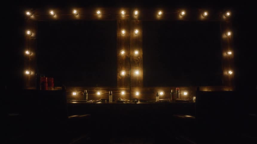 A double makeup vanity with classic theatre lights slowly illuminates a dark backstage dressing room