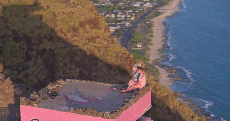 An aerial view reveals a romantic couple sitting on the edge of a unique pink structure on a mountainside, enjoying the vast ocean view at sunrise