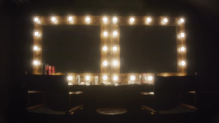A dark dressing room with two empty chairs in front of a brightly lit makeup vanity mirror slowly blurs out of focus