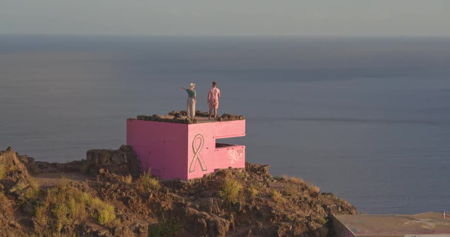 An aerial shot orbits a couple admiring the view from a pink breast cancer awareness bunker on a cliff overlooking the Hawaiian coastline and a rainbow
