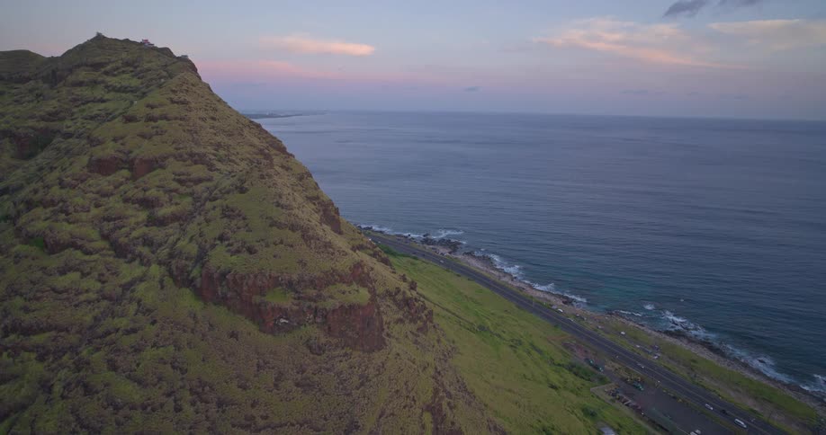 An epic aerial pull-back reveals a lush green mountain overlooking a scenic coastal highway and the vast ocean at twilight