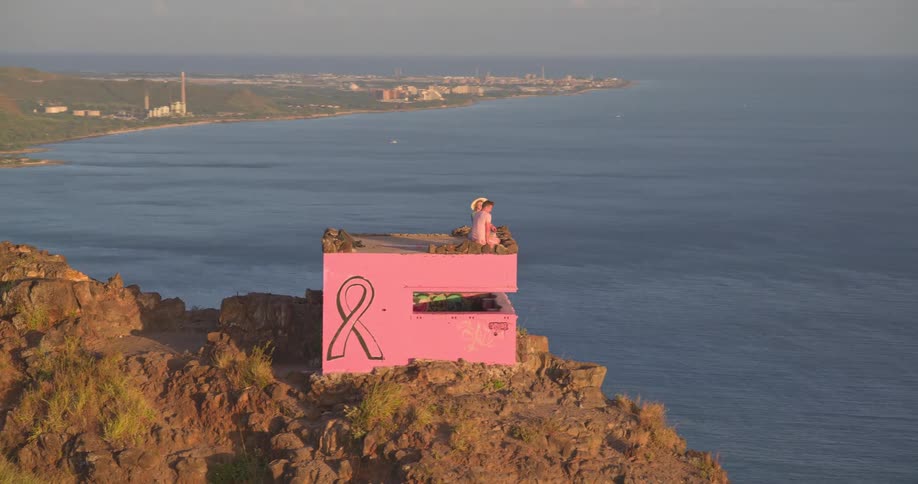 An aerial shot orbits a couple enjoying the scenic coastal view from a pink pillbox bunker with a breast cancer awareness ribbon painted on the side