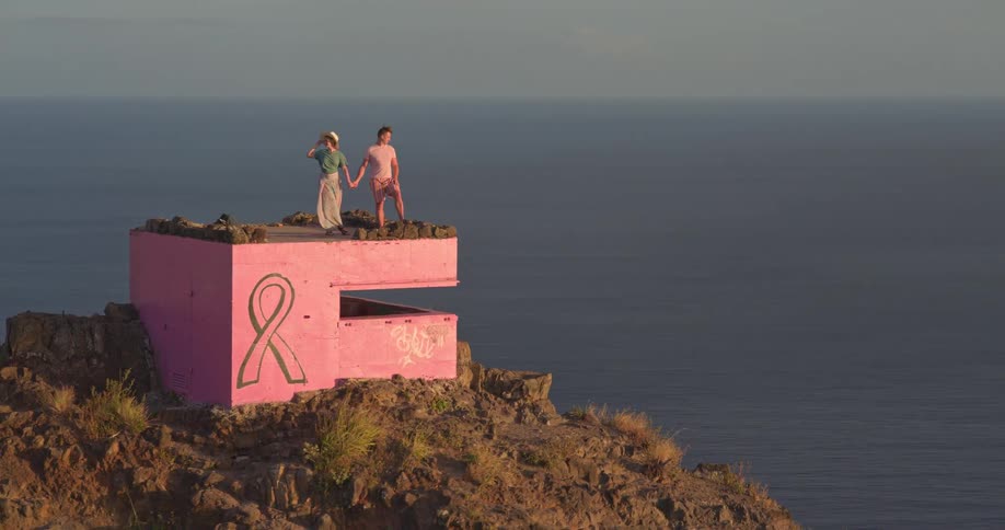 A romantic couple holds hands and enjoys the stunning ocean view from a pink bunker on a mountaintop in Hawaii at sunrise