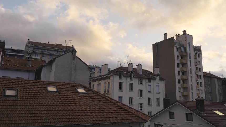 A serene view over the rooftops of a European city under a cloudy, golden-hour sky as birds fly past