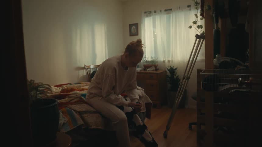 A woman with an injured leg sits on her bed and adjusts her medical brace, with crutches resting nearby in her bedroom