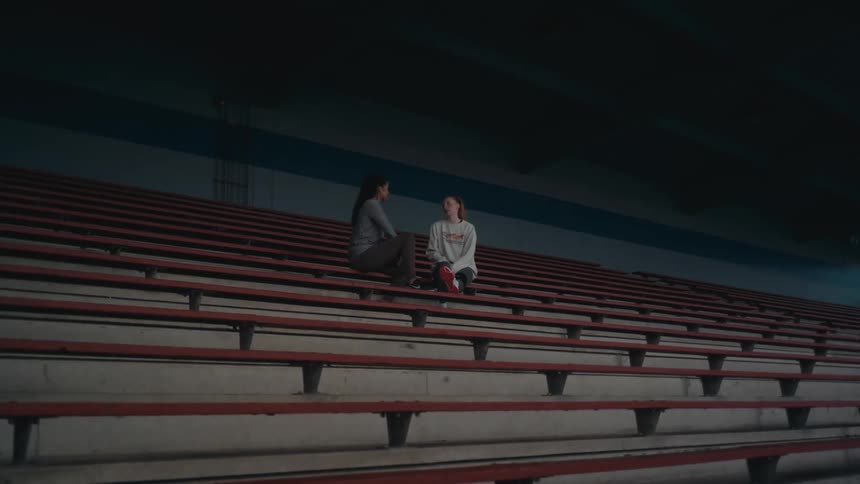 Two young women have a serious conversation while sitting alone on the bleachers in an empty, dimly lit arena