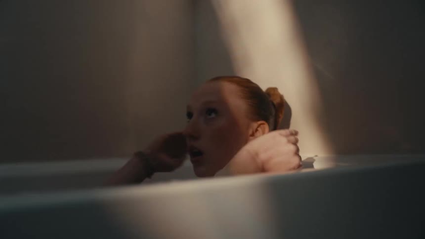 A distressed redhead sits in a bathtub, her face illuminated by a single ray of light as she looks up with an expression of fear