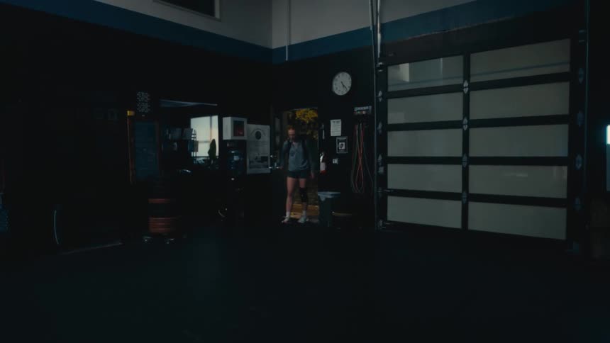 A woman in athletic wear and a knee brace walks into a dark, empty gym to begin her workout