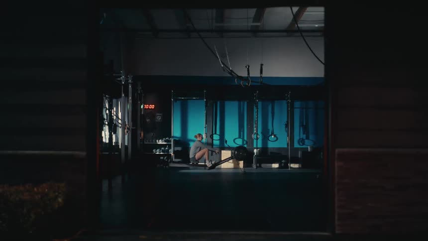 A lone female athlete trains on a rowing machine inside a dark, moody gym, viewed from outside