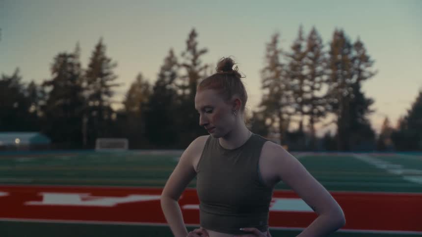 A determined young woman with red hair in a bun catches her breath while standing on a running track at dusk
