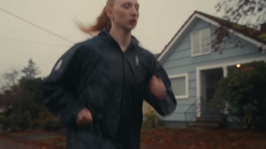An athletic woman with red hair runs in slow motion through a suburban neighborhood on an overcast day