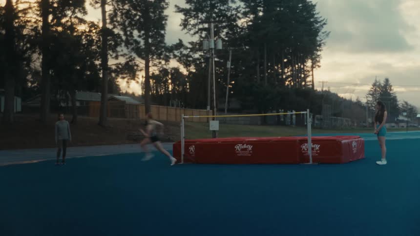 A track athlete runs towards the high jump bar for a practice attempt while two others watch on a blue track at sunset