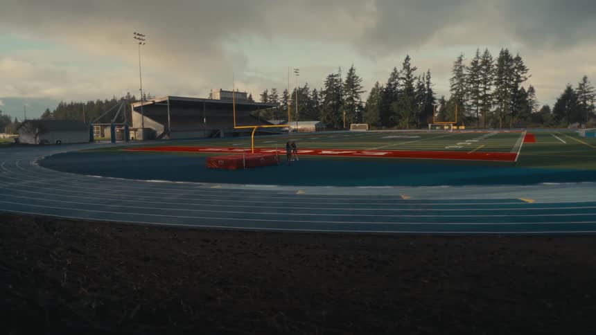 A wide, static shot captures two young women as they walk and talk on a blue running track at an empty stadium