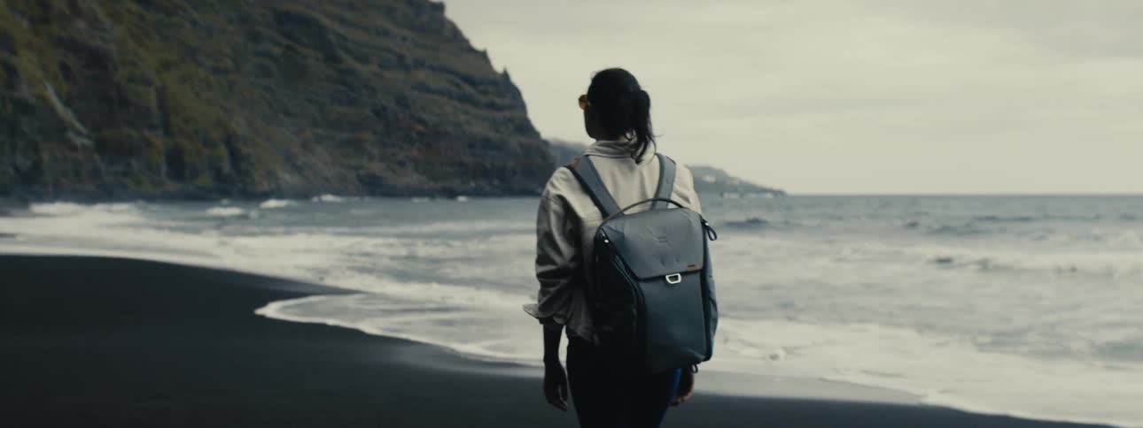 A woman with a backpack stands on a dark volcanic sand beach looking out at the ocean waves on an overcast day