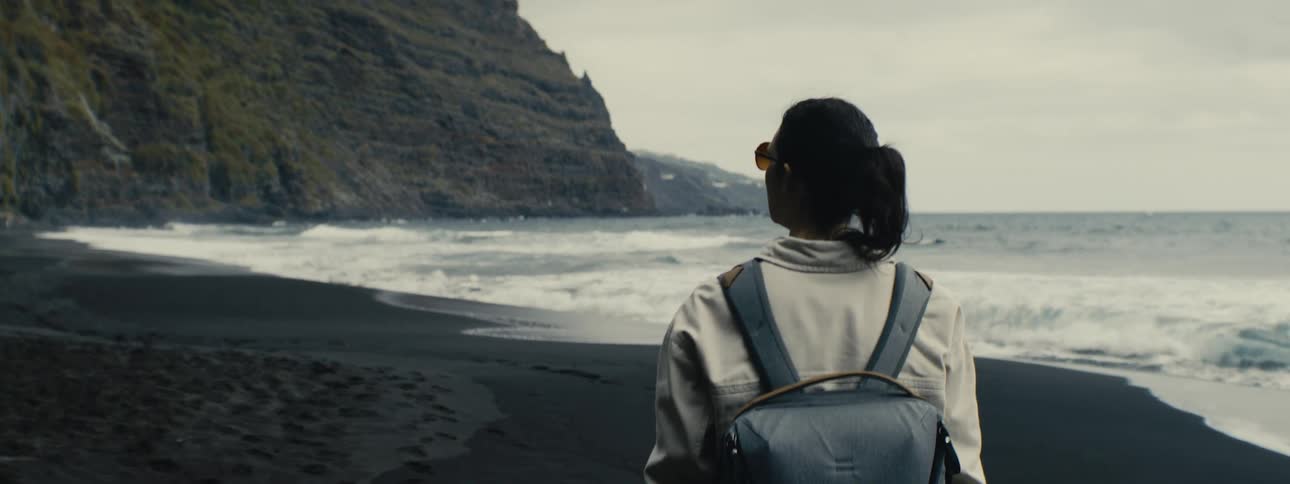 A lone traveler with a backpack stands on a black sand beach, watching the ocean waves crash against the shore
