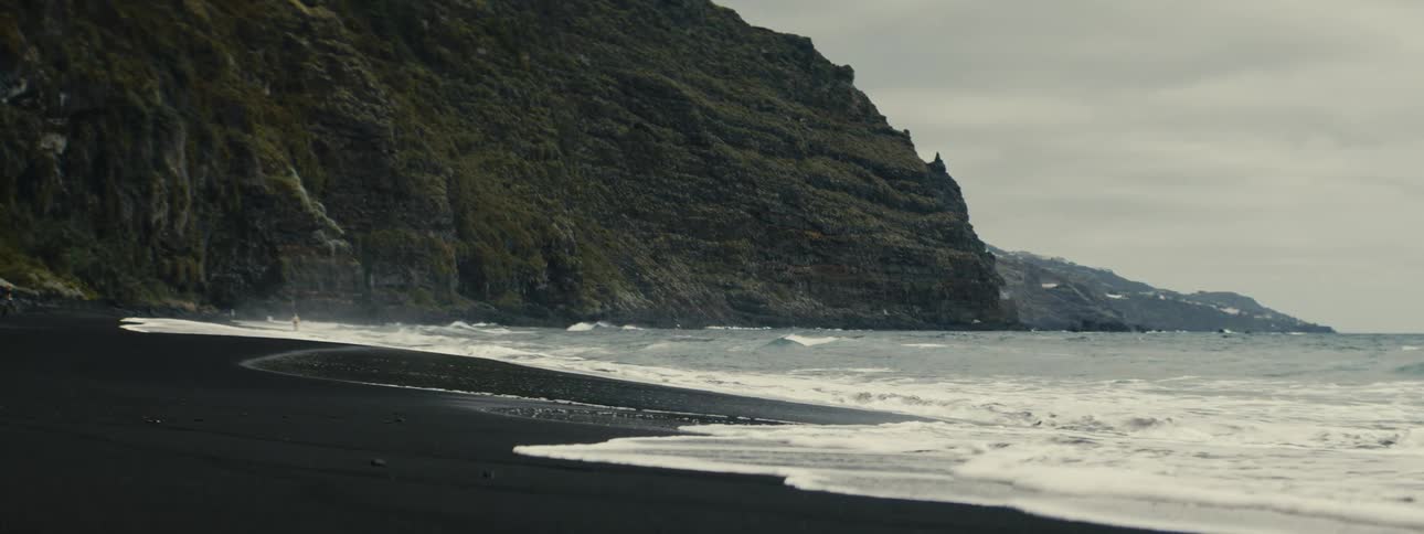 Slow motion waves wash ashore on a moody black sand beach at the base of a green cliff
