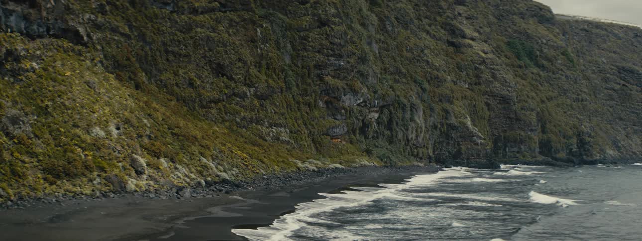 Waves gently roll onto a remote black sand beach nestled against a rugged, moss-covered cliff