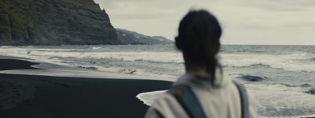 Slow motion shot from behind a woman as she watches the powerful ocean waves crash onto a black sand beach