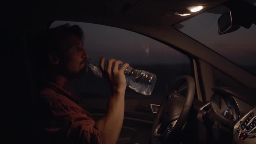 A man opens a bottle of water and drinks while sitting in a vehicle