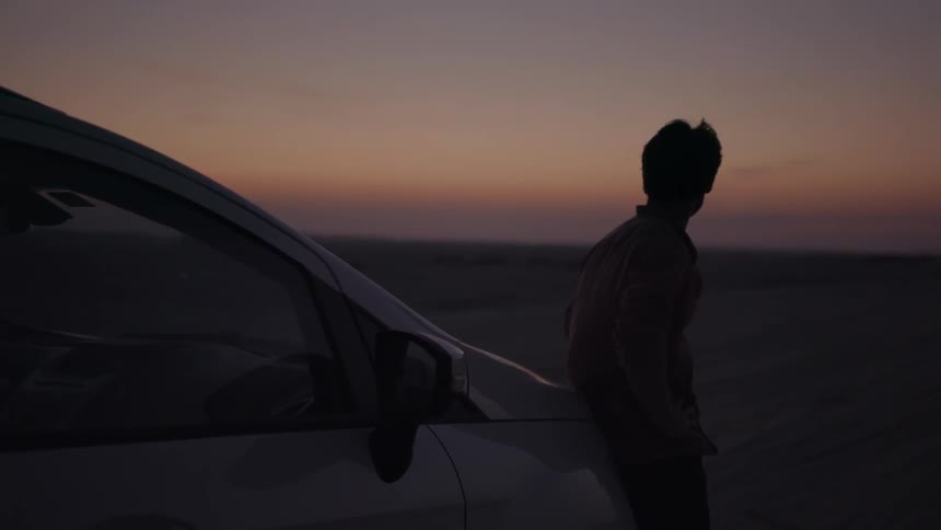 A man leans against his car at sunset looking at the scenery before he leaves