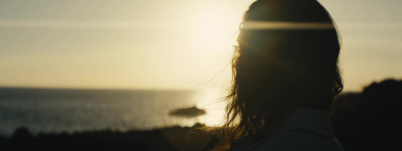 Close up of a silhouetted woman with windblown hair pensively watching a beautiful golden sunset over the ocean