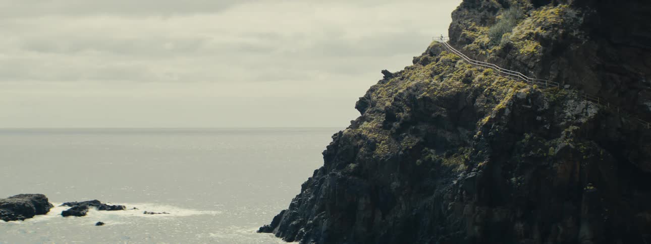 A solitary figure walks along a railed path atop a rugged, green-topped cliff overlooking the vast, calm sea