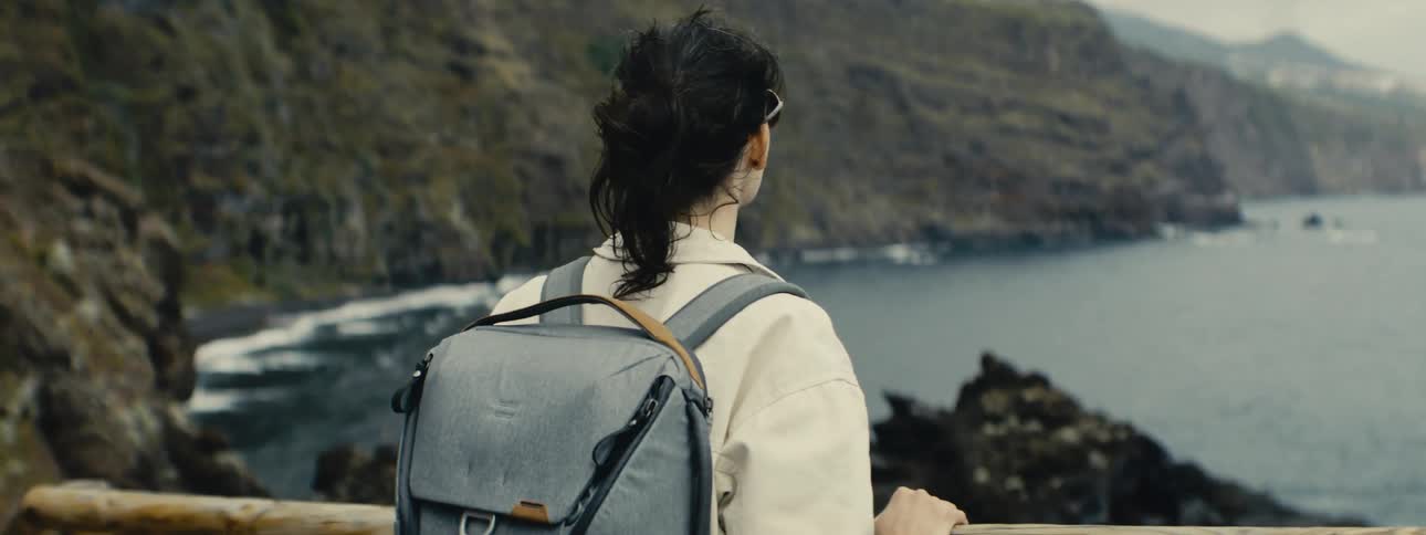 A woman with a backpack stands at a coastal viewpoint, her hair blowing in the wind as she looks out over the ocean in slow motion