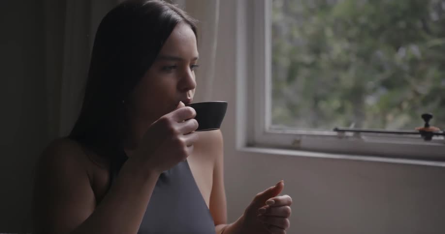 A woman sips from a steaming teacup while sitting by a window