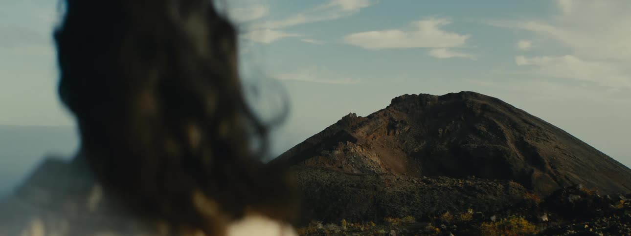 Over-the-shoulder shot of a person with long hair blowing in the wind, gazing at a large volcanic mountain at golden hour