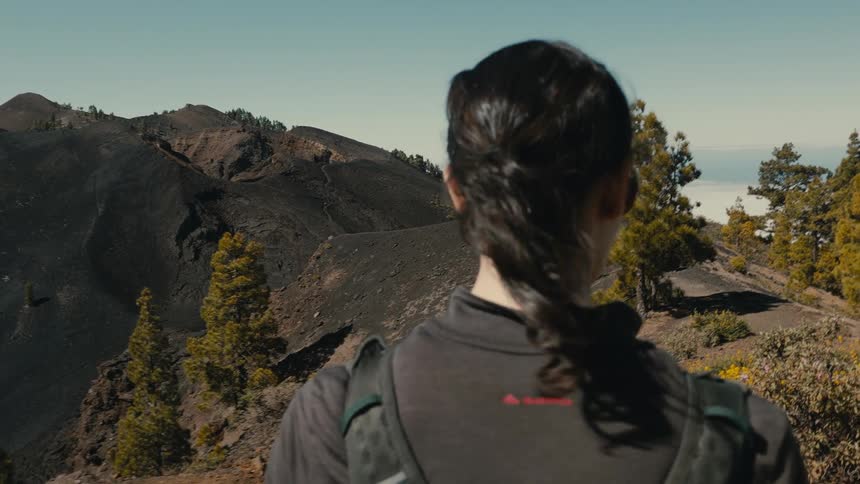 A woman with a backpack gazes at a breathtaking volcanic landscape in slow motion, her hair gently blowing in the wind