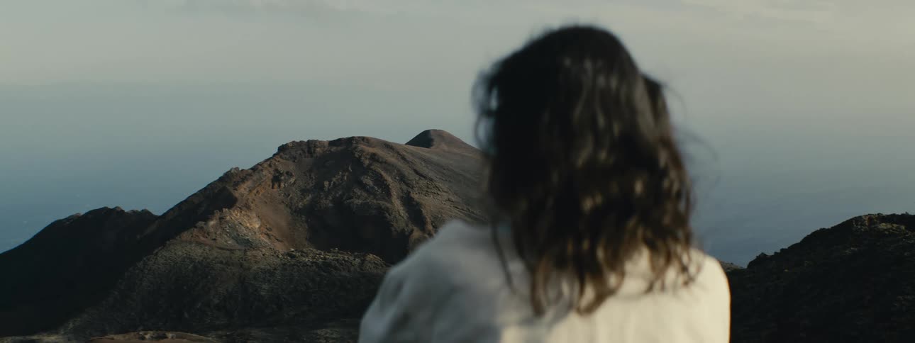 A rack focus shot from a person's back to a large volcanic mountain peak in a vast, serene landscape