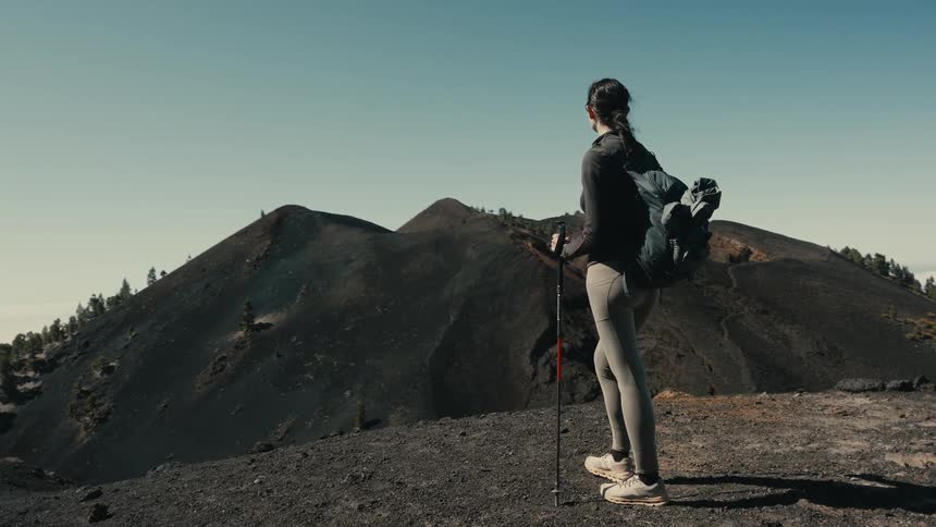 A female hiker with a backpack and trekking poles stands on a volcanic summit, taking in the vast and dramatic landscape