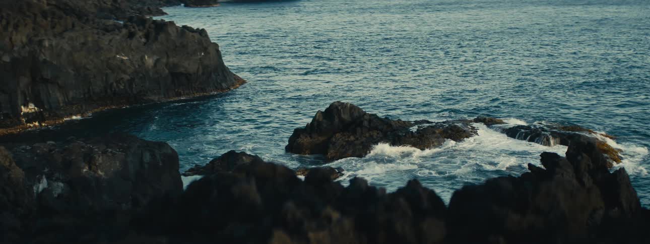 Gentle ocean waves crash against a rugged, dark volcanic rock shoreline in a serene and peaceful scene