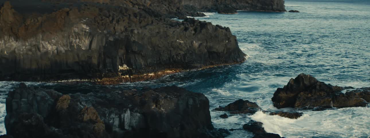 Powerful ocean waves crash in slow motion against a dramatic and rugged volcanic rock coastline at dusk