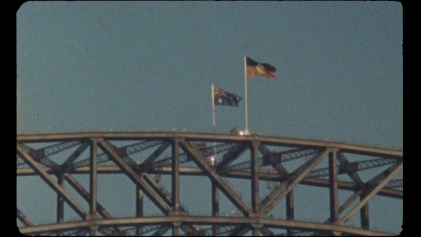 A vintage film shot shows the Australian and Aboriginal flags flying together atop the Sydney Harbour Bridge against a clear blue sky