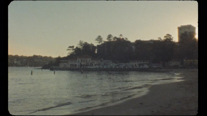 Vintage super 8 film captures a tranquil view of a calm bay with gentle waves and distant shoreline buildings at sunset